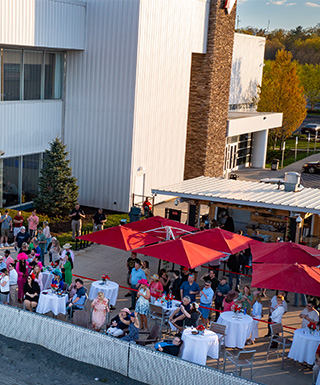 Aerial view of guests enjoying an outdoor event on the trackside patio at Plainridge Park Casino, featuring red umbrellas, set tables, and the racetrack fence, with the casino building in the background.